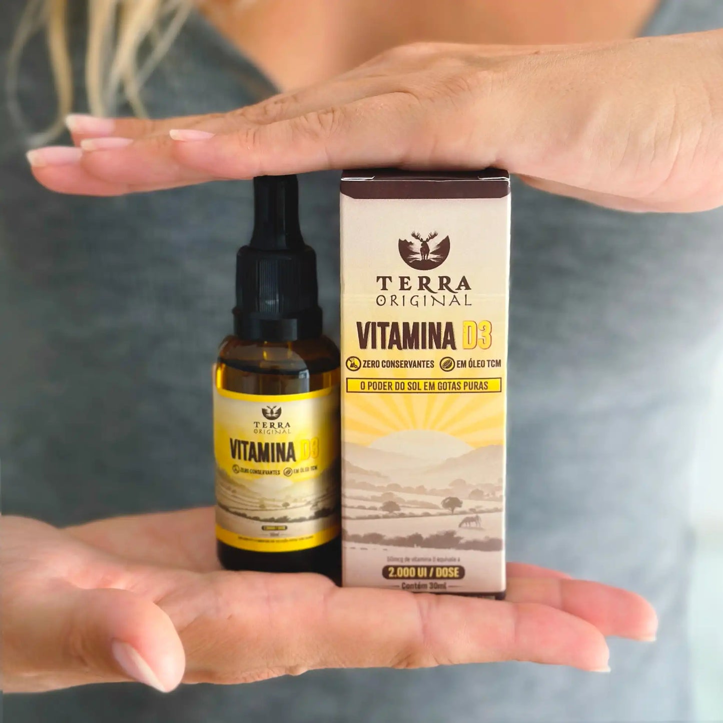Pessoa exibindo frasco e caixa de vitamina D3 em óleo TCM Terra Original apoiados na mão com fundo neutro