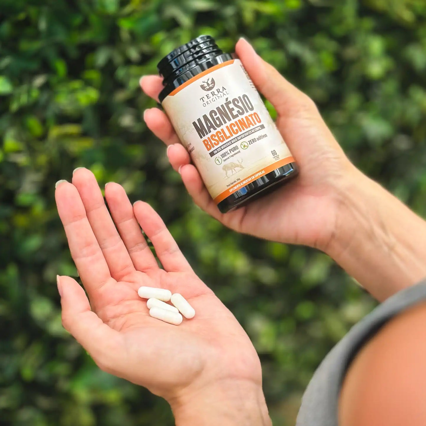 Mão segurando frasco de magnésio bisglicinato Terra Original e outra mão mostrando cápsulas brancas do suplemento em ambiente externo